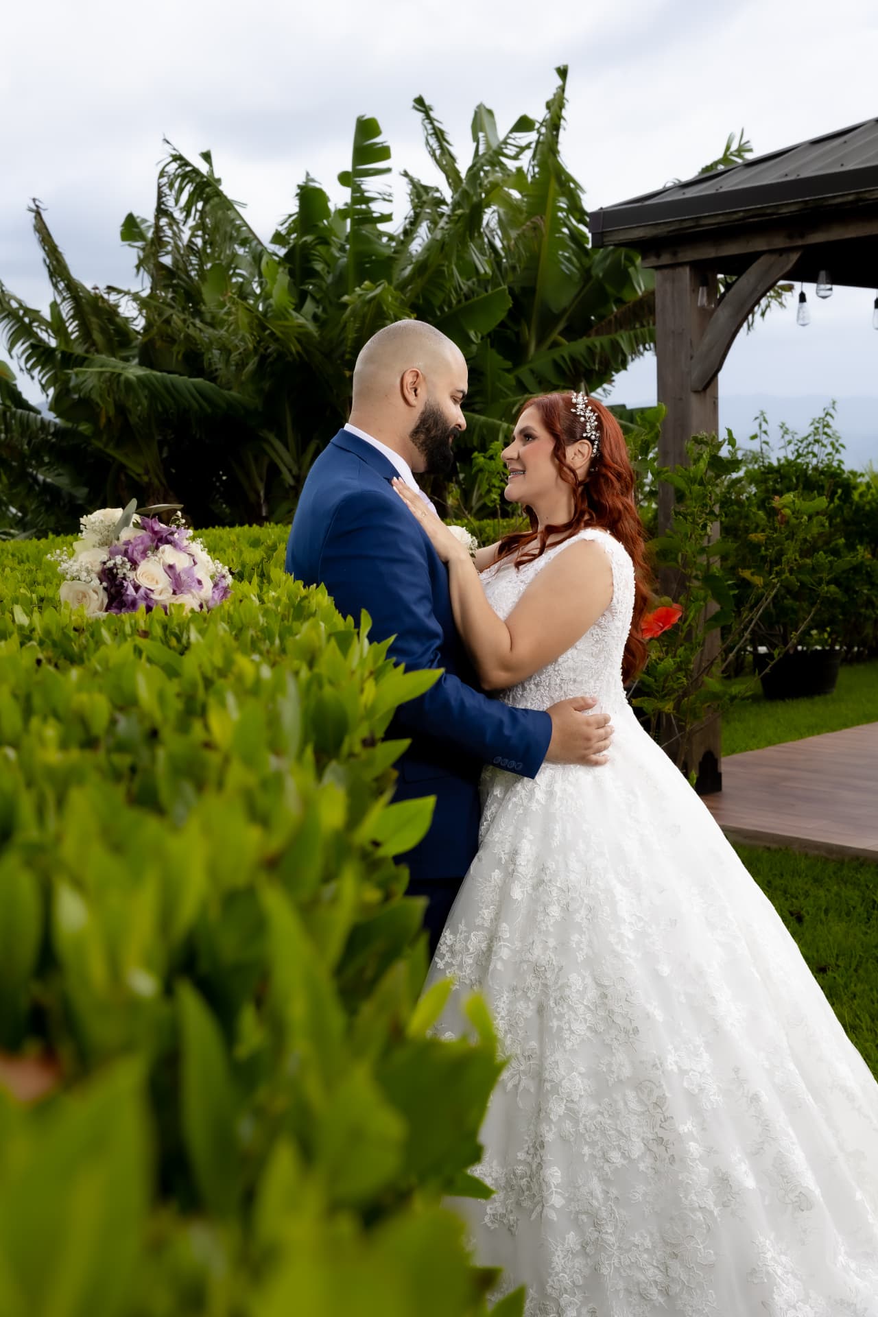 Pareja de novios abrazada entre jardín tropical y pérgola de madera