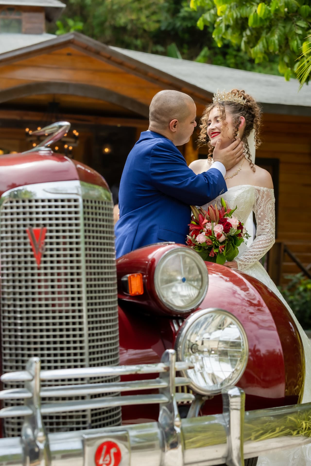 Pareja de novios junto a un auto clásico rojo frente a una hacienda de madera