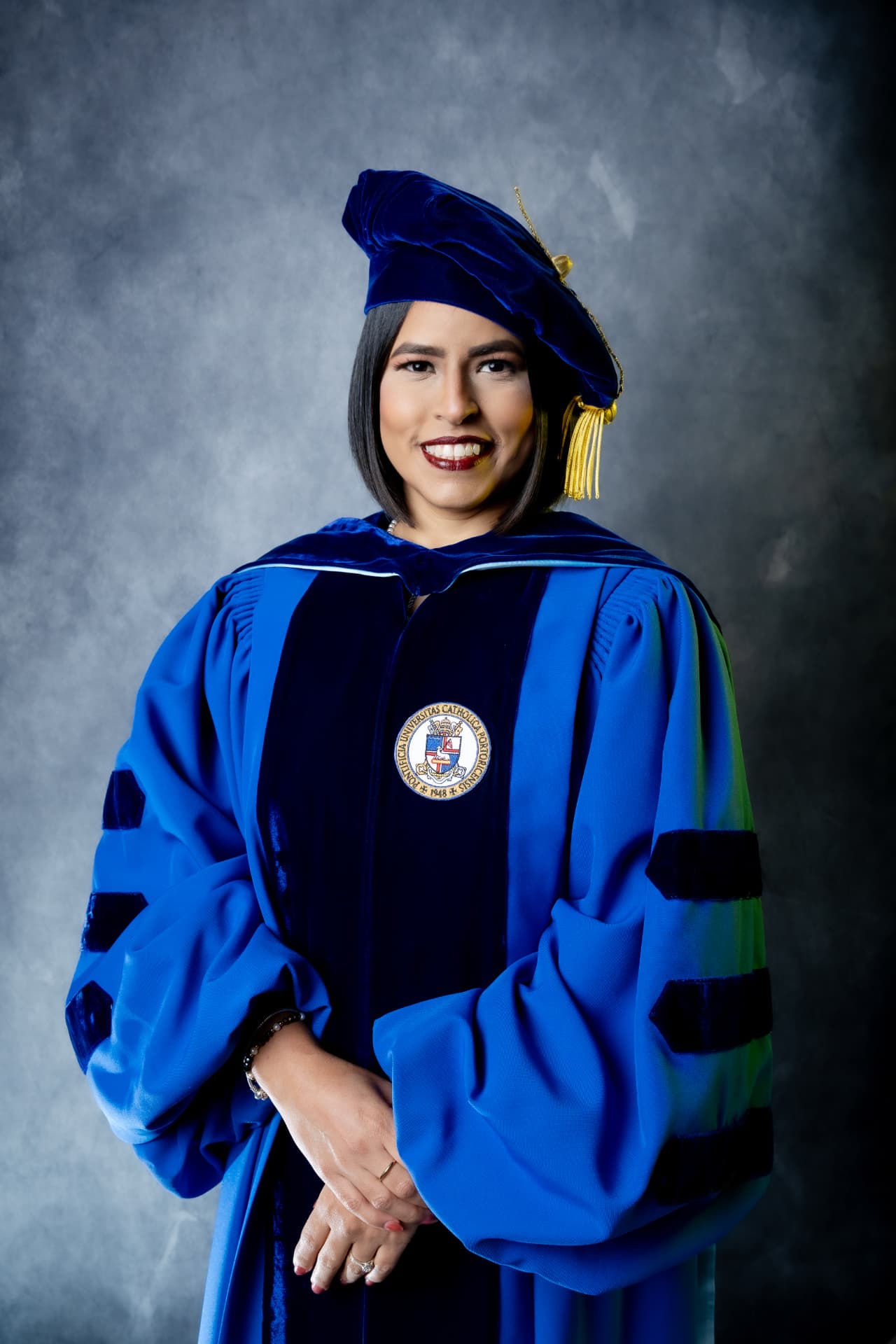Graduanda con toga doctoral azul y estola dorada sobre fondo gris
