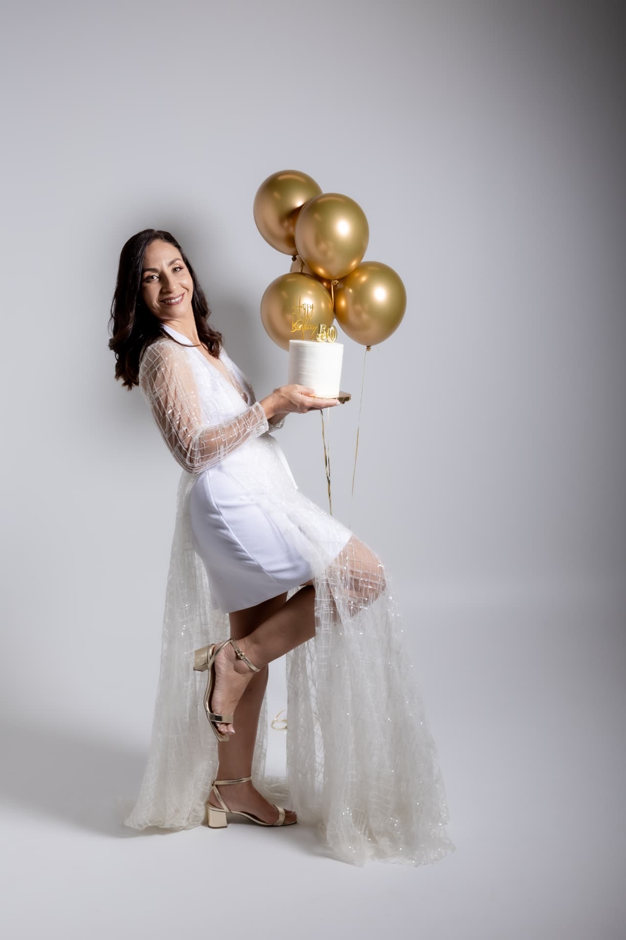 Mujer con vestido blanco sosteniendo pastel y globos dorados