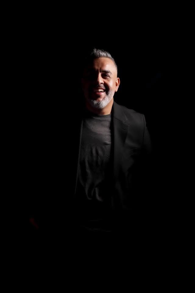 Hombre con barba canosa y saco negro sobre fondo oscuro