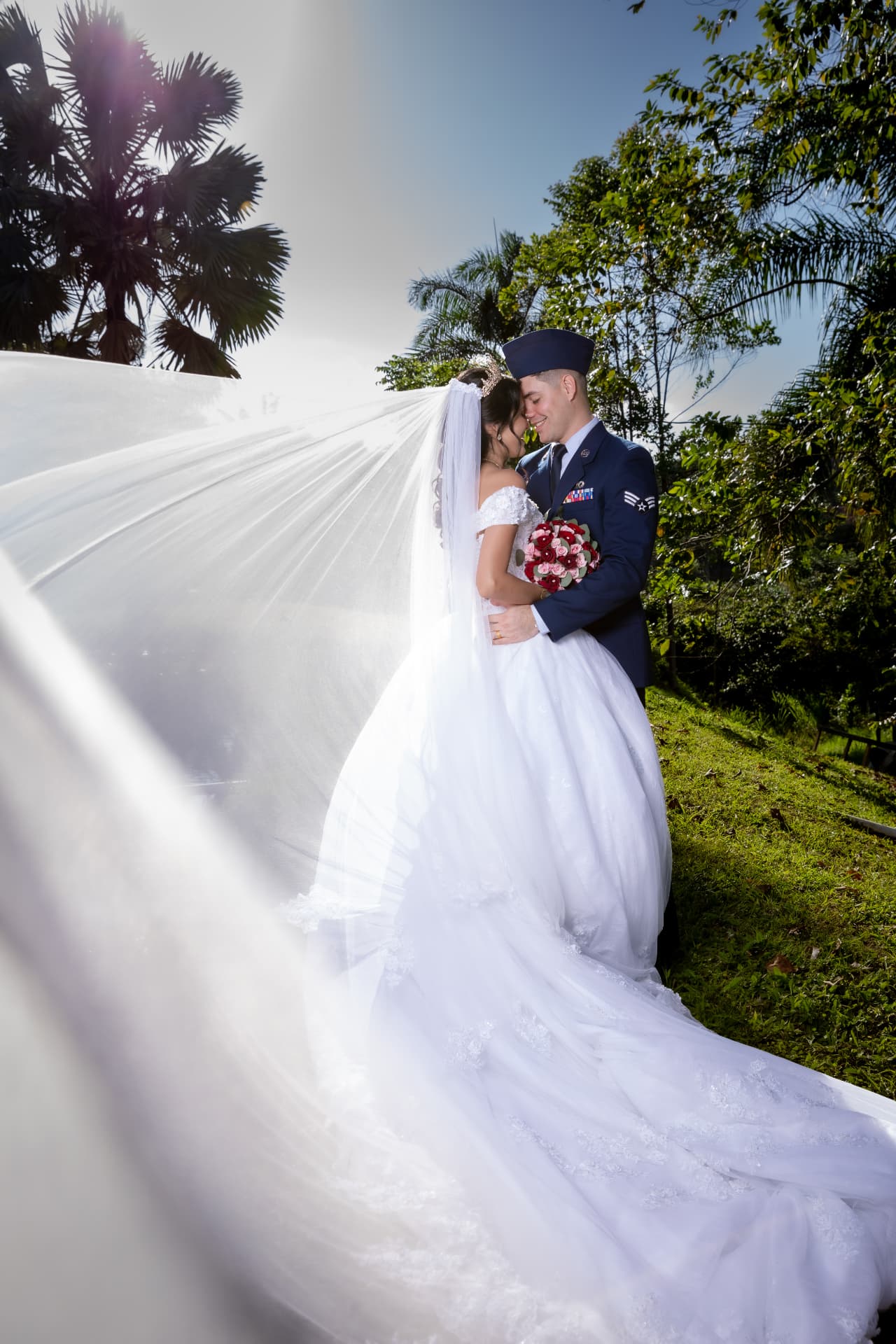 Novia con velo largo junto a su pareja en uniforme militar bajo palmeras