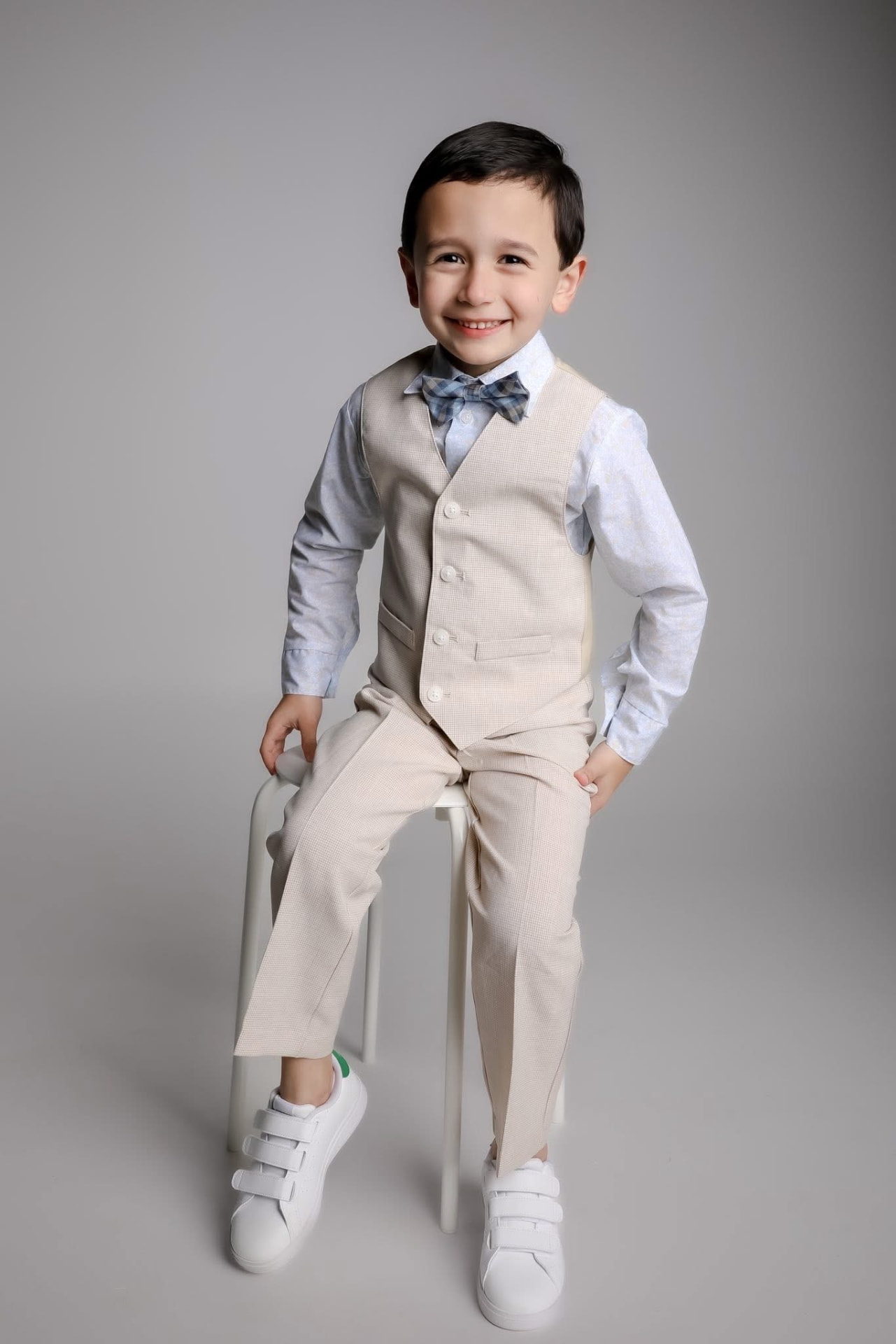 Niño sonriente con traje beige y moño celeste sentado en taburete
