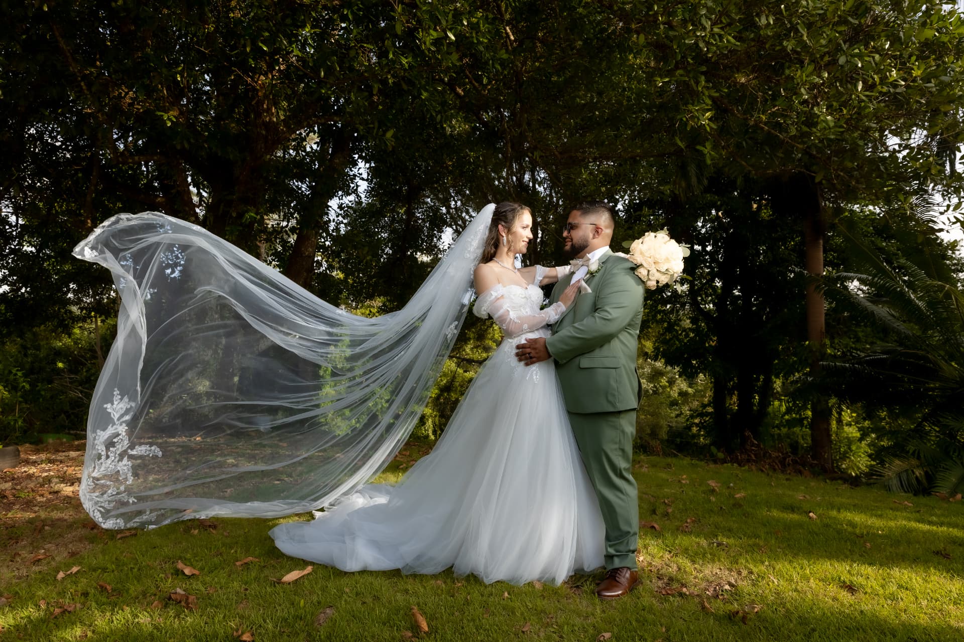 Novia con velo largo fluyendo junto al novio en jardín verde